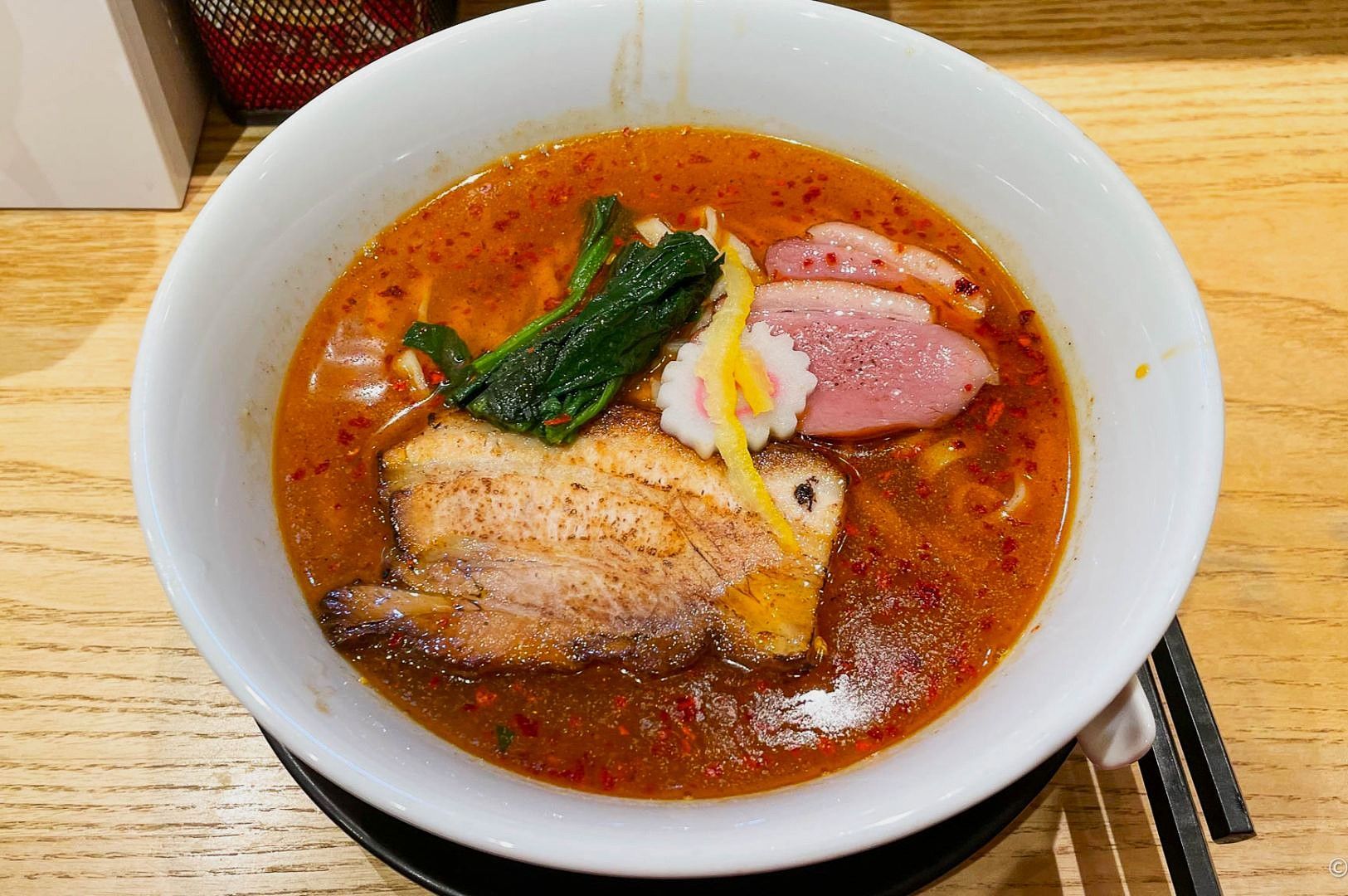 A white ceramic ramen bowl sits on a wooden table, filled with a spicy orange broth and crowned by thick slices of chashu pork, wilted greens, a pink-and-white narutomaki, and a bright yellow garnish.