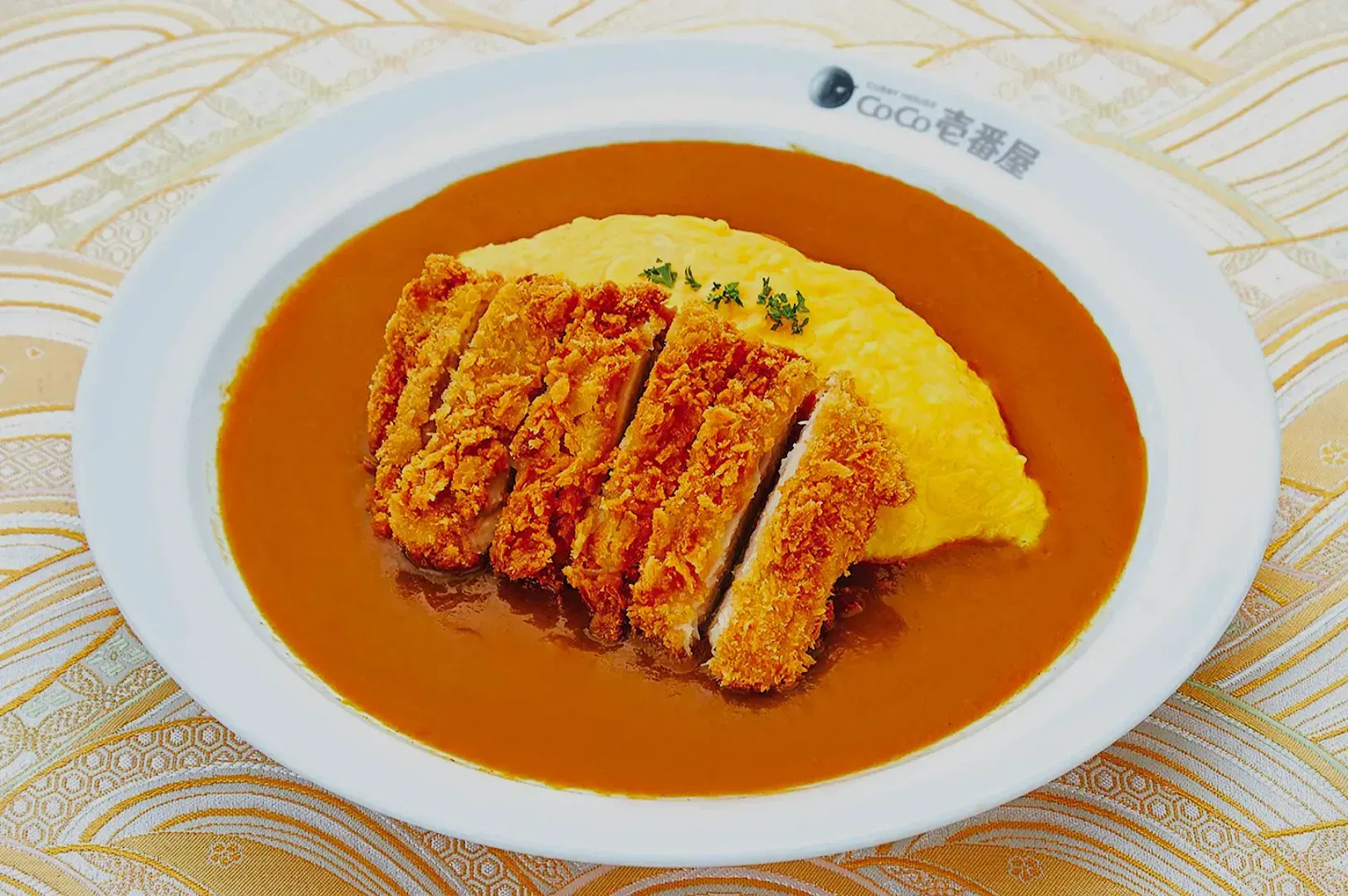 A plate of Japanese curry features a golden breaded cutlet and a fluffy omelet, garnished with herbs, on a patterned background. Comforting and appetizing.