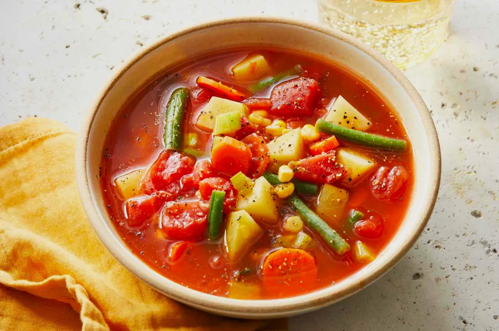 A vibrant bowl of mixed vegetable soup, featuring tomatoes, potatoes, carrots, and green beans in a rich broth. A yellow napkin adds warmth.