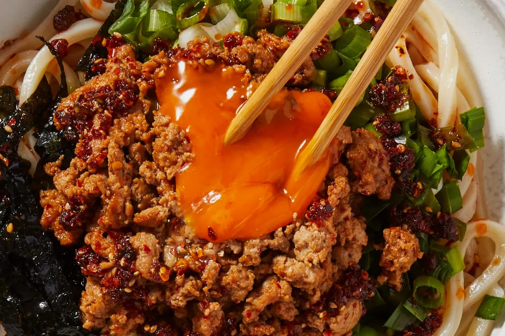 A bowl of noodles topped with spicy minced meat, a broken egg yolk, chopped green onions, and seaweed slices, with chopsticks poised above.
