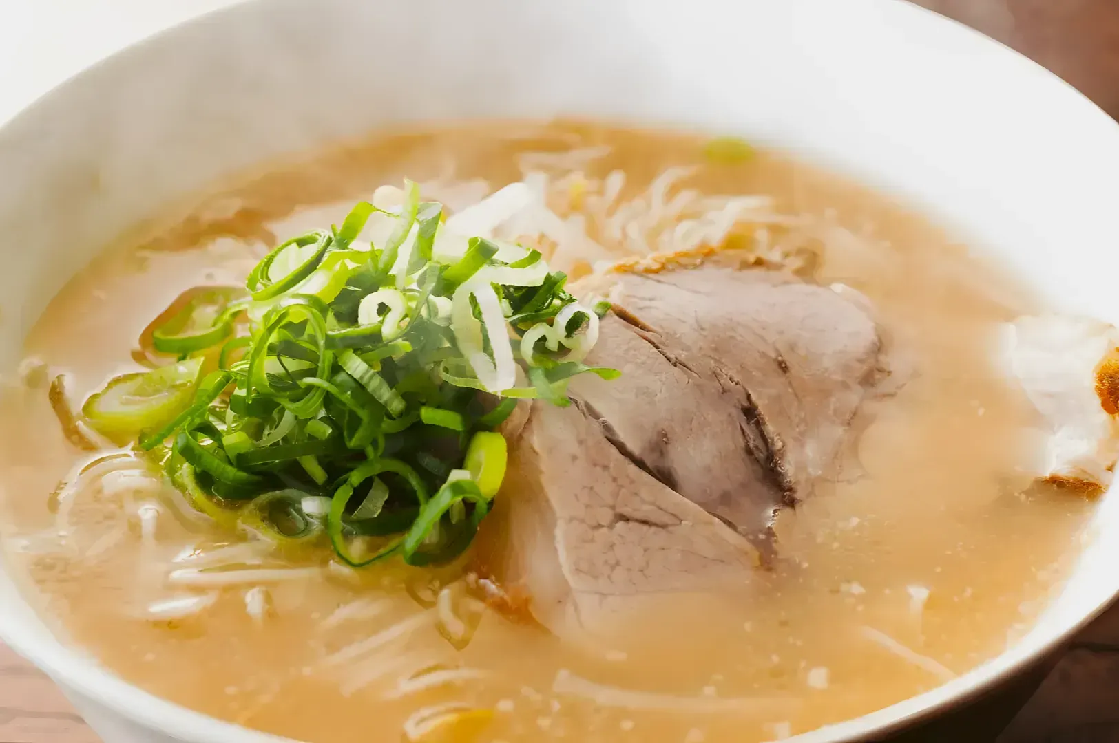 A steaming bowl of ramen topped with slices of pork, chopped green onions, and white noodles in a rich, light broth on a wooden table.