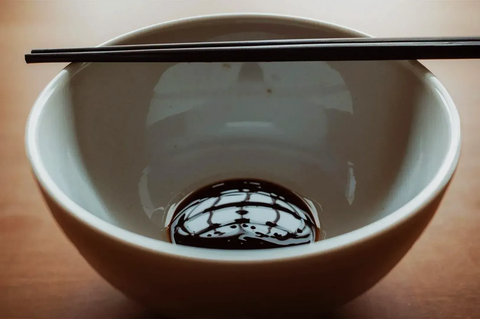 A white bowl with a small amount of dark soy sauce at the bottom, reflecting light. Two chopsticks rest on top. The scene conveys simplicity and calm.