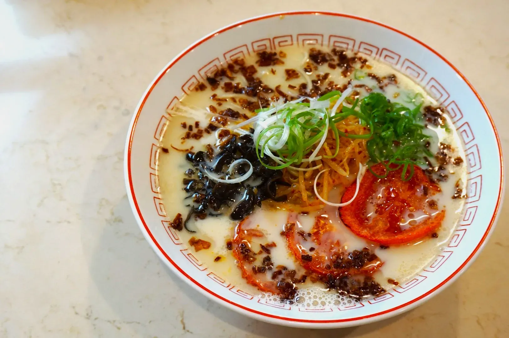 A bowl of creamy ramen with sliced green onions, tomatoes, black mushrooms and crispy toppings on a marble surface, conveying warmth and richness.