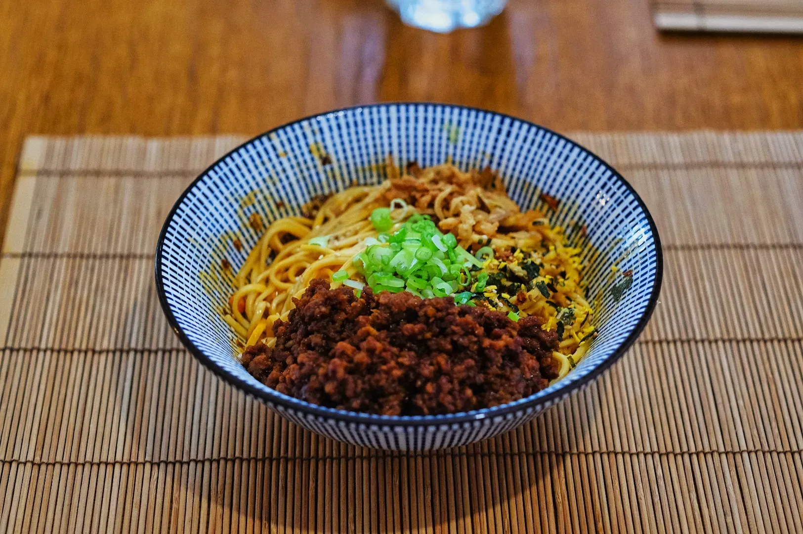 A striped bowl of noodles topped with minced meat, green onions, and crispy bits sits on a bamboo mat, creating a warm and inviting presentation.