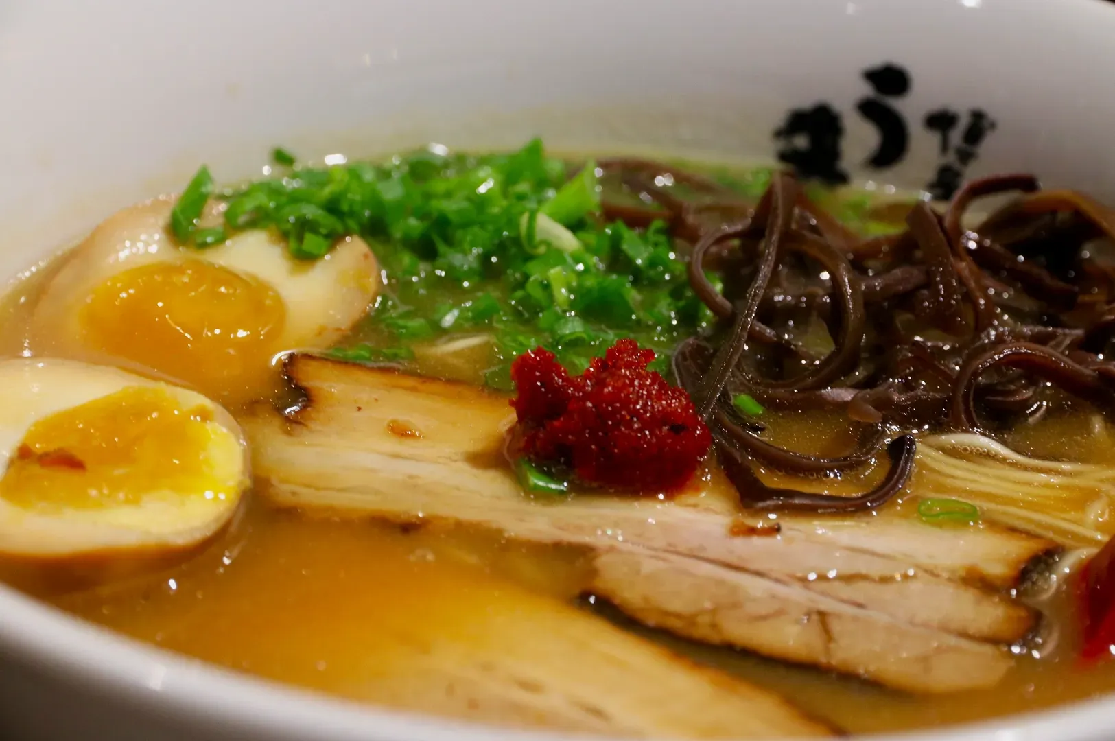 Bowl of ramen with sliced pork, soft-boiled egg halves, chopped green onions, mushrooms, and a dollop of red paste, in rich broth.