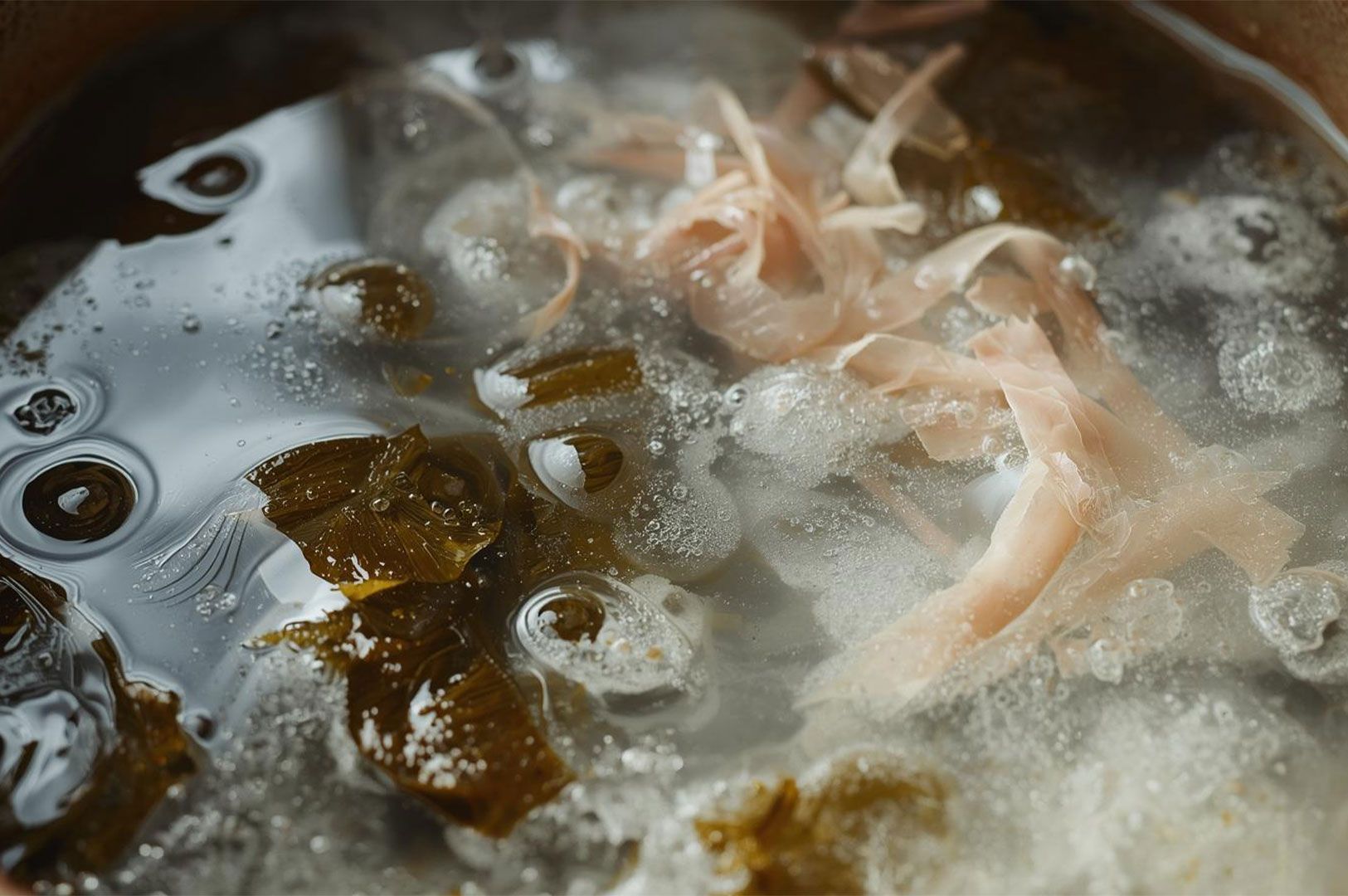 A close-up view of clear, simmering dashi (broth) with pieces of dark green kelp (kombu) and delicate, pinkish-brown bonito flakes floating on the surface.
