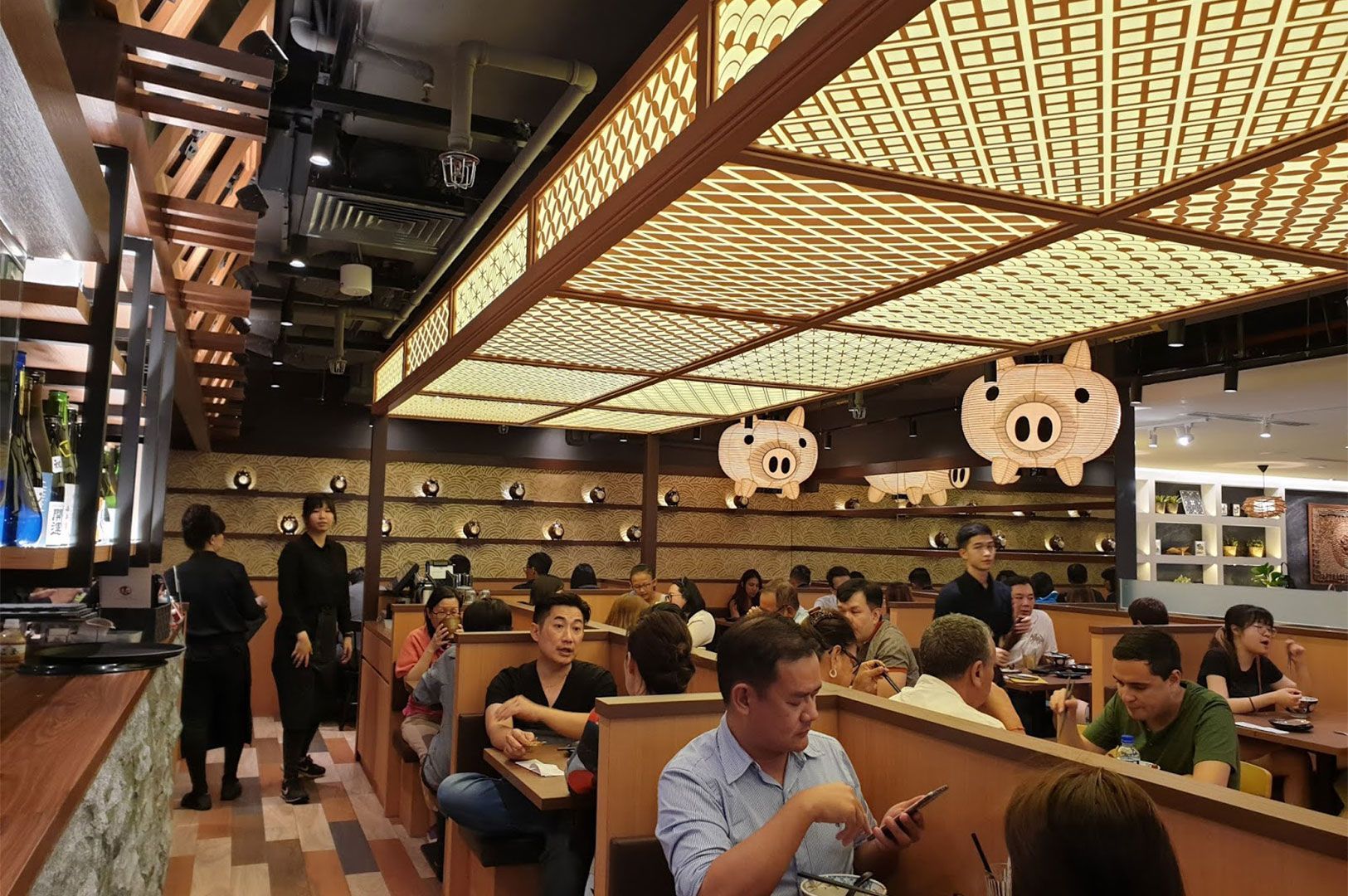 A busy interior shot of a Japanese ramen restaurant with a low, textured, wooden ceiling. Diners are seated at tables under unique, light-up pig-shaped lanterns hanging from the ceiling.