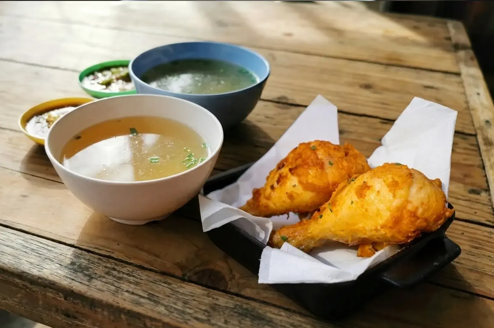 Golden fried chicken drumsticks served with clear chicken broth soup and dipping sauces on rustic wooden table.