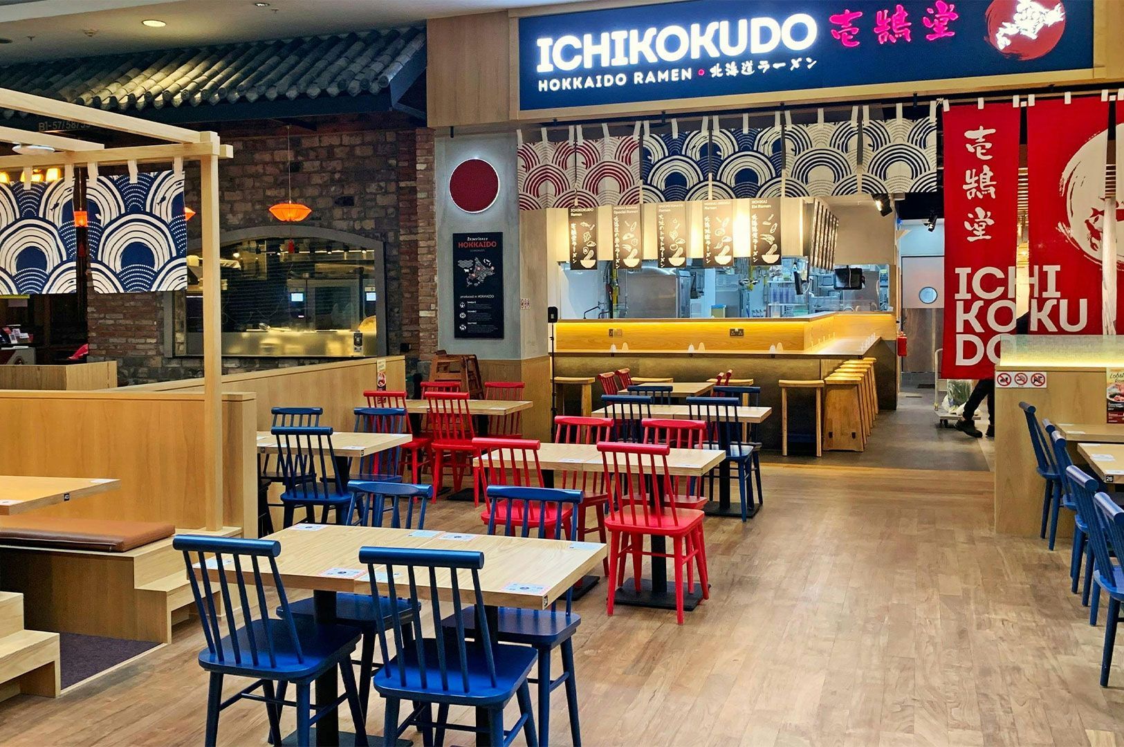 Interior view of an Ichikokudo Hokkaido Ramen restaurant, featuring Japanese-style decor with red and blue chairs, wooden tables, and an open kitchen counter.