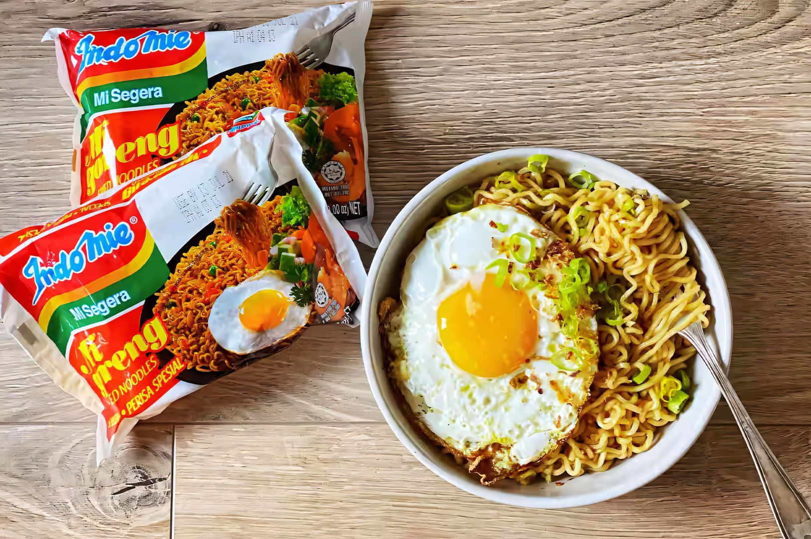 Overhead shot of a bowl of Indomie Mi Goreng instant noodles topped with a sunny-side-up fried egg and sliced green onions, next to two unopened packets of the noodles.