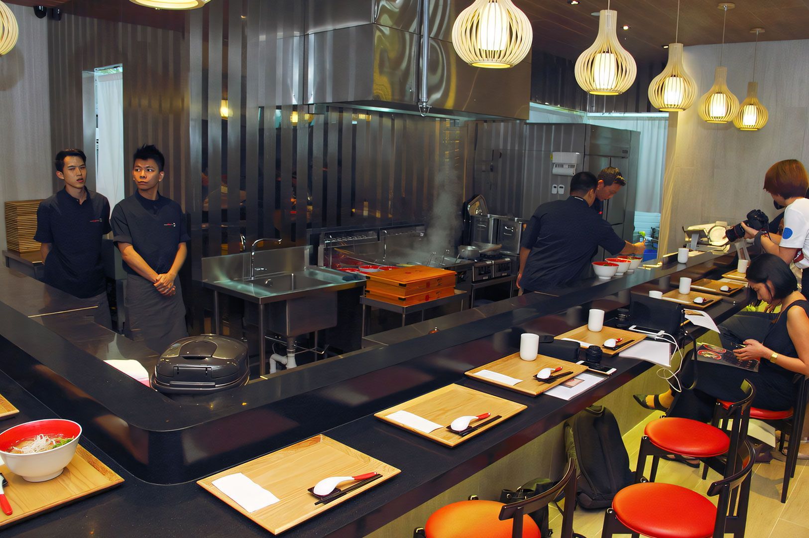 Contemporary Japanese restaurant with an open kitchen, black counter seating, orange bar stools, and distinctive modern hanging pendant lights.