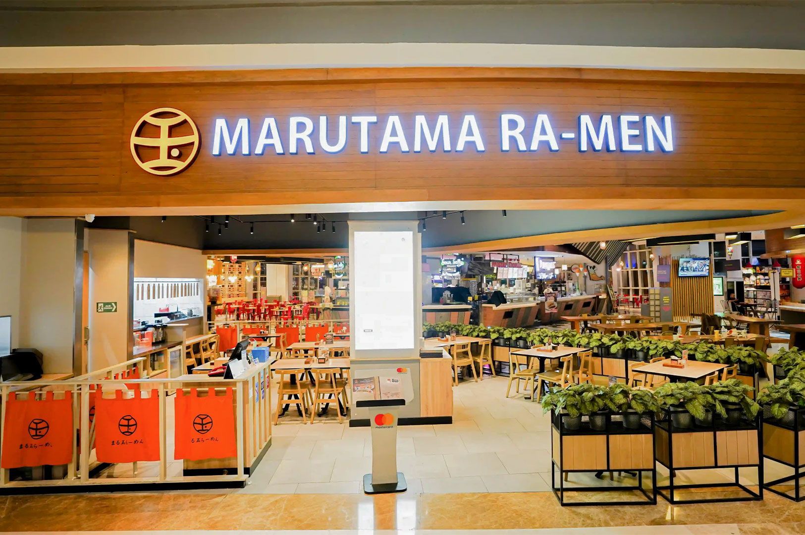Storefront view of a Marutama Ra-Men restaurant in a mall setting, featuring its logo on a wooden sign and a bright, open interior with tables and potted plants.