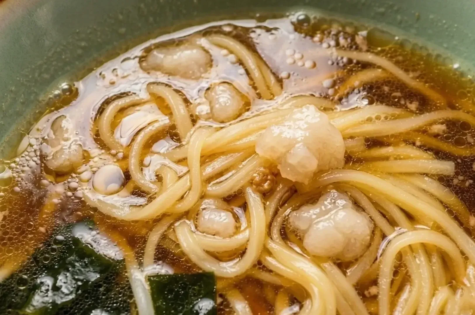 Close-up of flat ramen noodles in clear dashi broth with pork fat and seaweed.