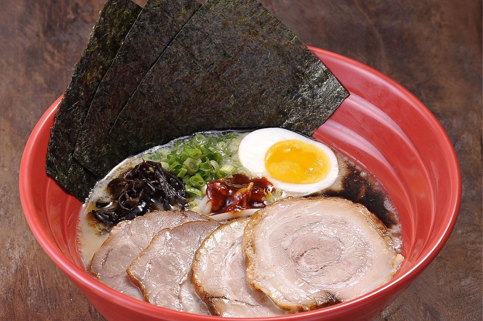 Bowl of spicy ramen with thick slices of chashu pork, half a soft-boiled egg, black fungus, chili oil, and three large sheets of nori seaweed, served in a red bowl.