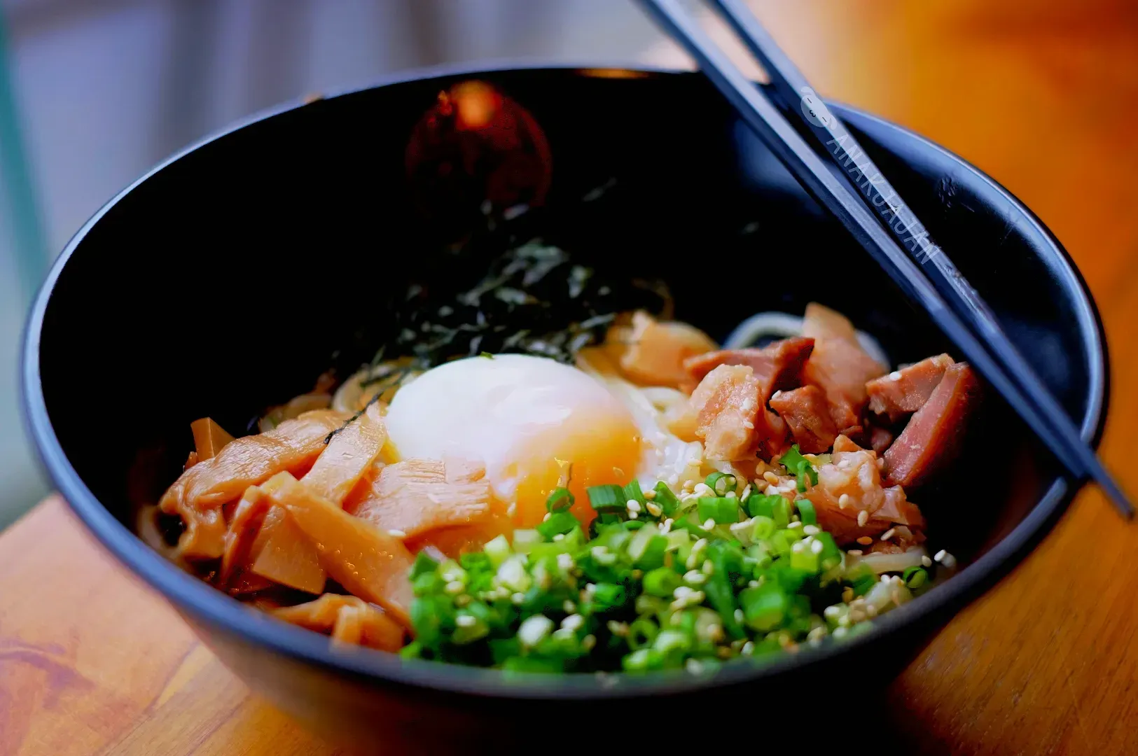 A black bowl of ramen with sliced bamboo shoots, green onions, seaweed, tender pork, and a soft-boiled egg, with chopsticks on the side.