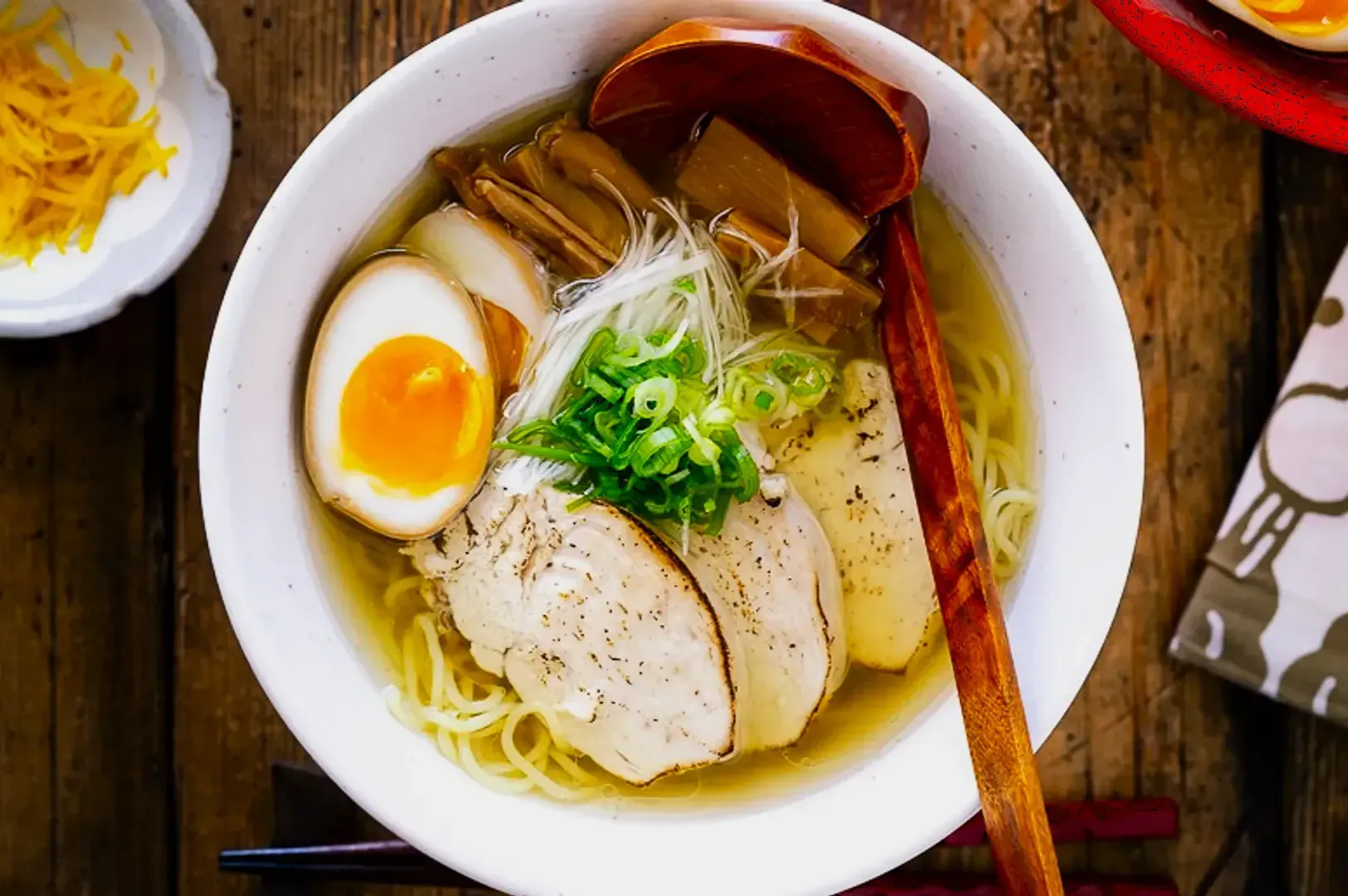 A bowl of chicken ramen with clear broth, featuring sliced chicken, a halved soft-boiled egg, scallions, and a wooden spoon, set on a rustic wooden table.