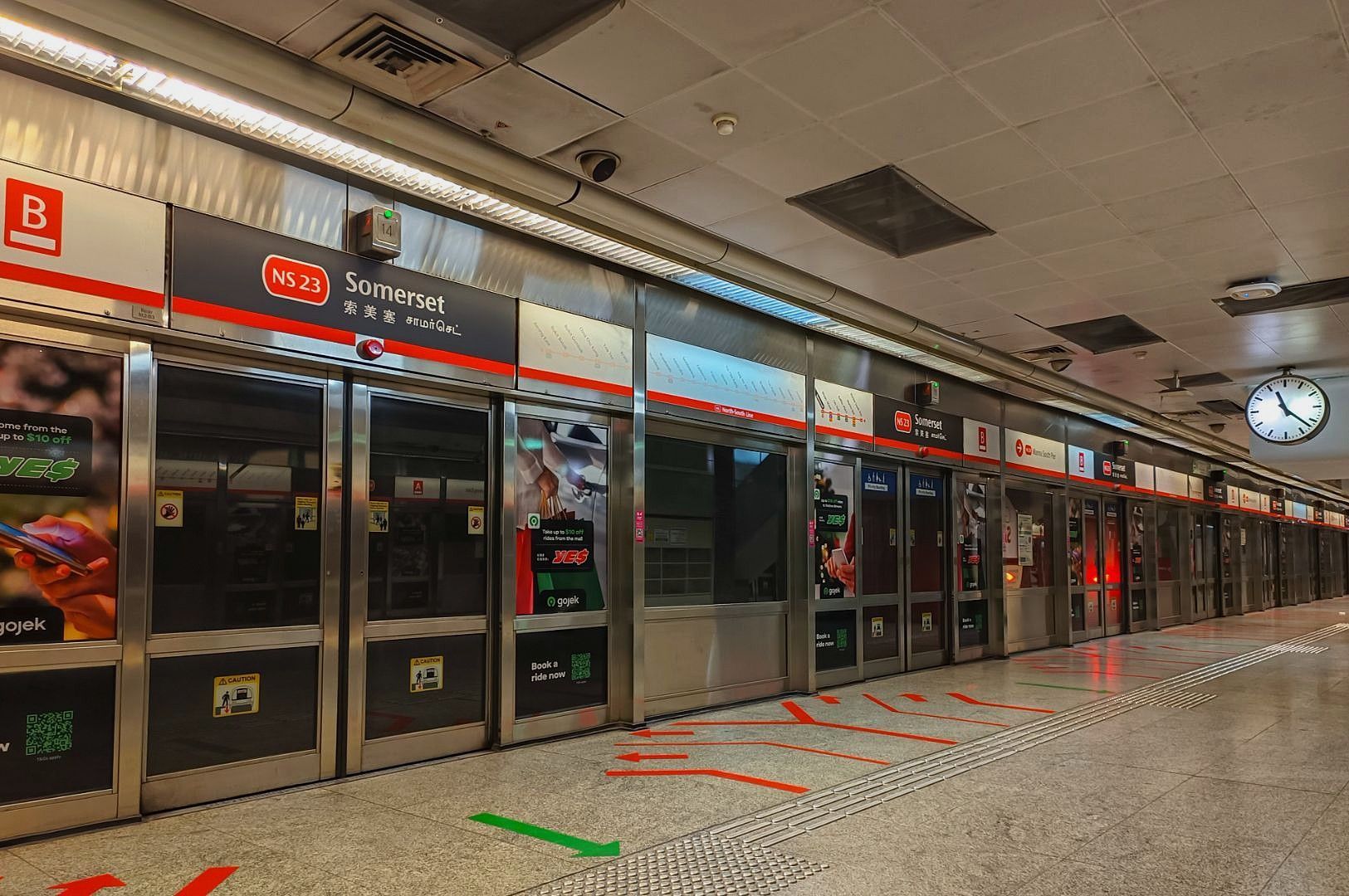 Empty subway platform at Somerset Station, featuring closed glass doors with service signs above. Bright lighting, a clock on the wall, and directional floor markings.