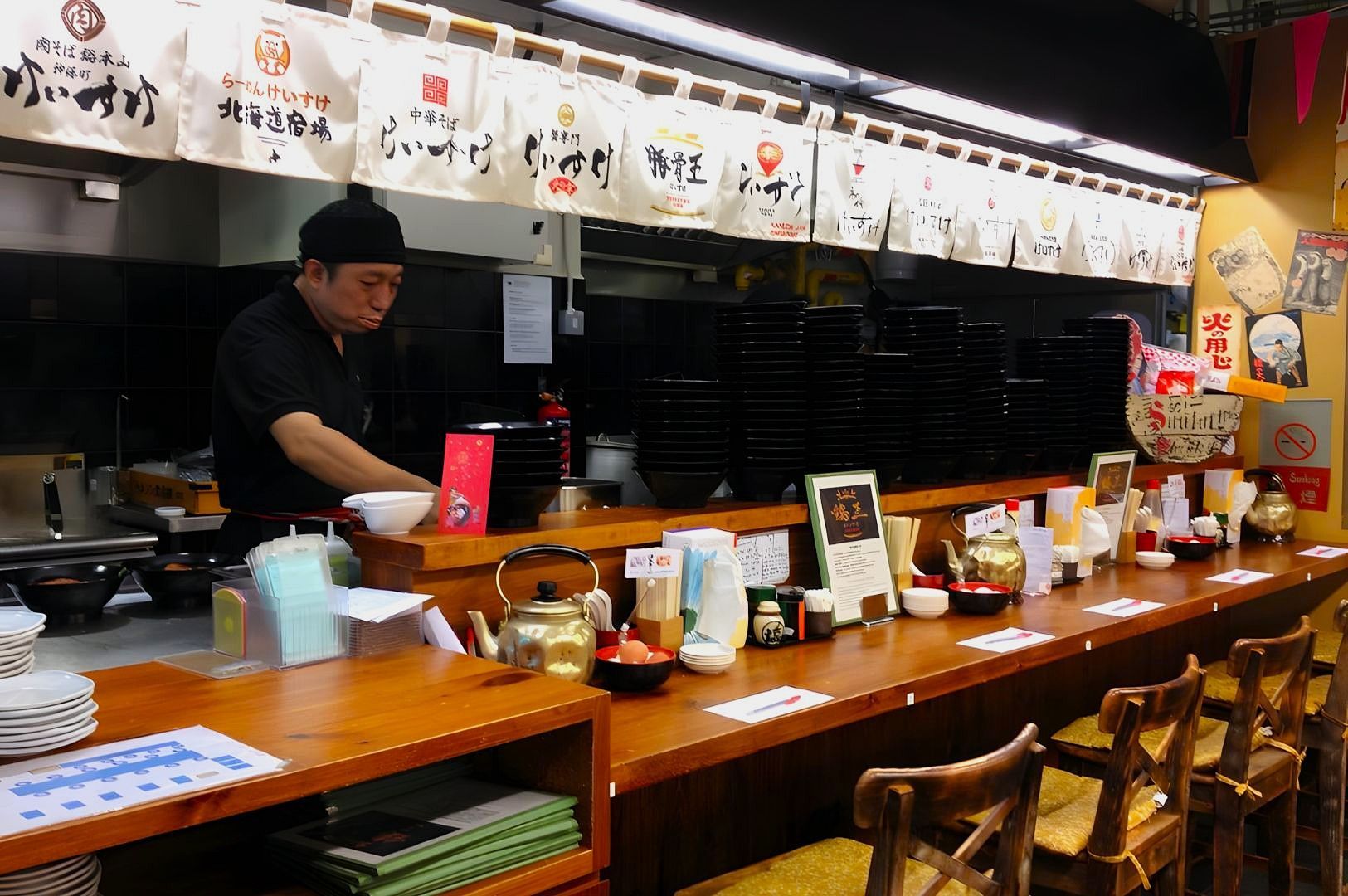 A professional chef in a black uniform and cap stands behind a wooden ramen counter, preparing bowls while a row of tall black bowls stacks up behind him under white fabric banners.