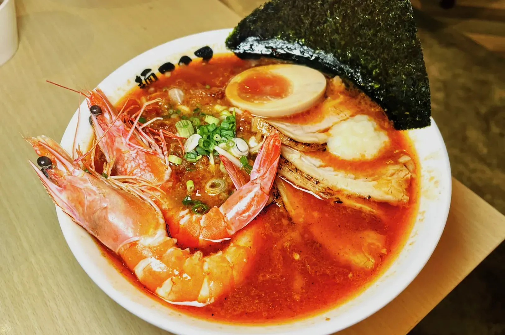 A steaming bowl of ramen featuring vibrant shrimp, a soft-boiled egg, sliced pork, green onions, nori, and rich red broth, creating a savory and inviting dish.
