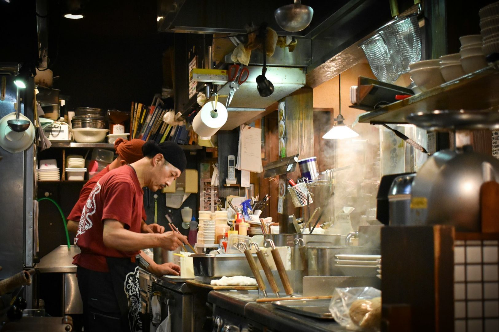 A focused chef in a red shirt prepares food in a bustling, dimly lit kitchen filled with steam, utensils, and a cluttered array of equipment and dishes.