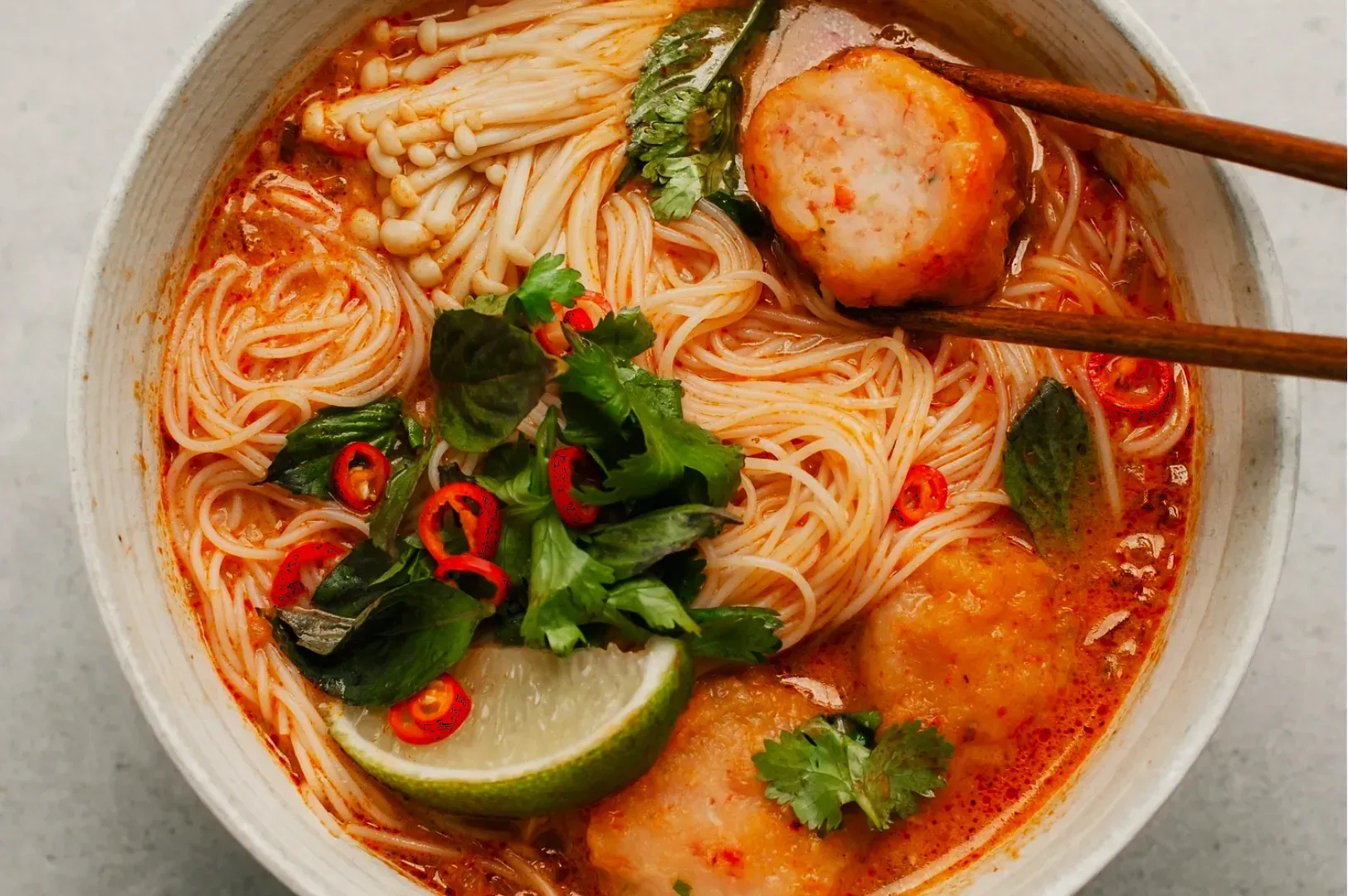 A bowl of spicy ramen soup with thin noodles, fish balls, and mushrooms, garnished with fresh herbs, red chili slices, and a lime wedge. Chopsticks hold a fish ball, conveying a savory and vibrant meal.
