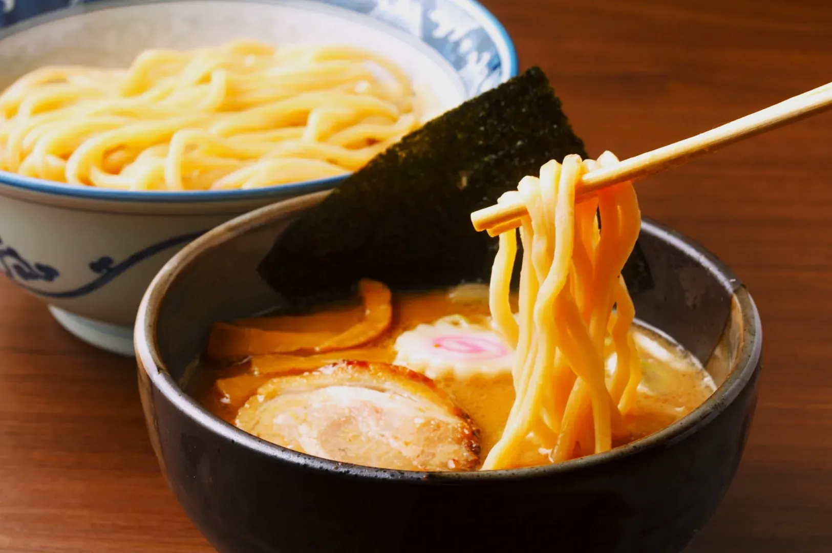 Close-up of a steaming bowl of tsukemen noodles; thick noodles are dipped into rich broth with slices of pork, narutomaki, and seaweed. Cozy and appetizing.