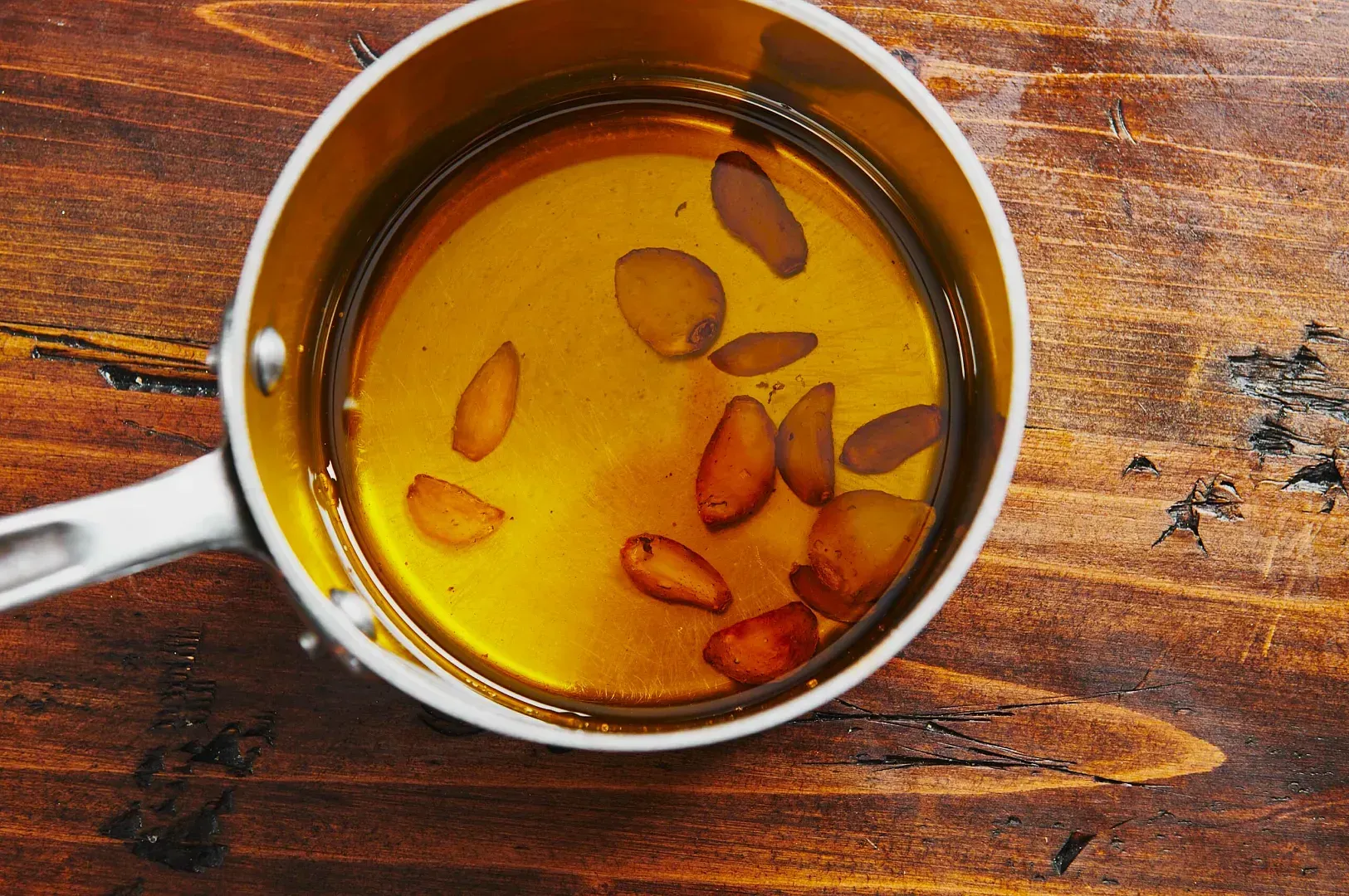 A small metal pot filled with golden oil and slices of browned garlic on a rustic wooden table. The scene conveys a warm, inviting culinary setting.