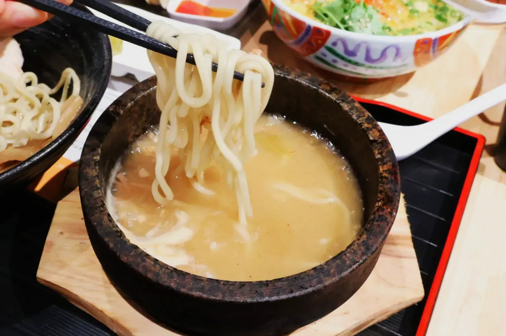 Chopsticks lift thick noodles from a stone bowl filled with steaming broth, suggesting a hearty meal. In the background, a colorful bowl adds a vibrant touch.