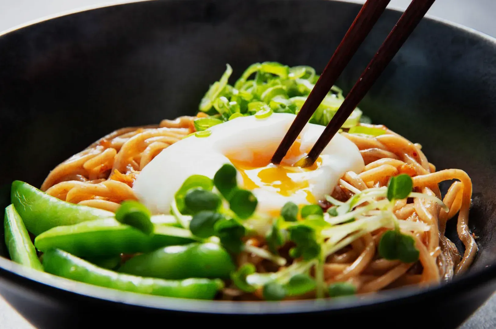 A black bowl of noodles topped with snow peas, green onions, and a soft-boiled egg. Chopsticks pierce the egg, releasing the yolk over the noodles.