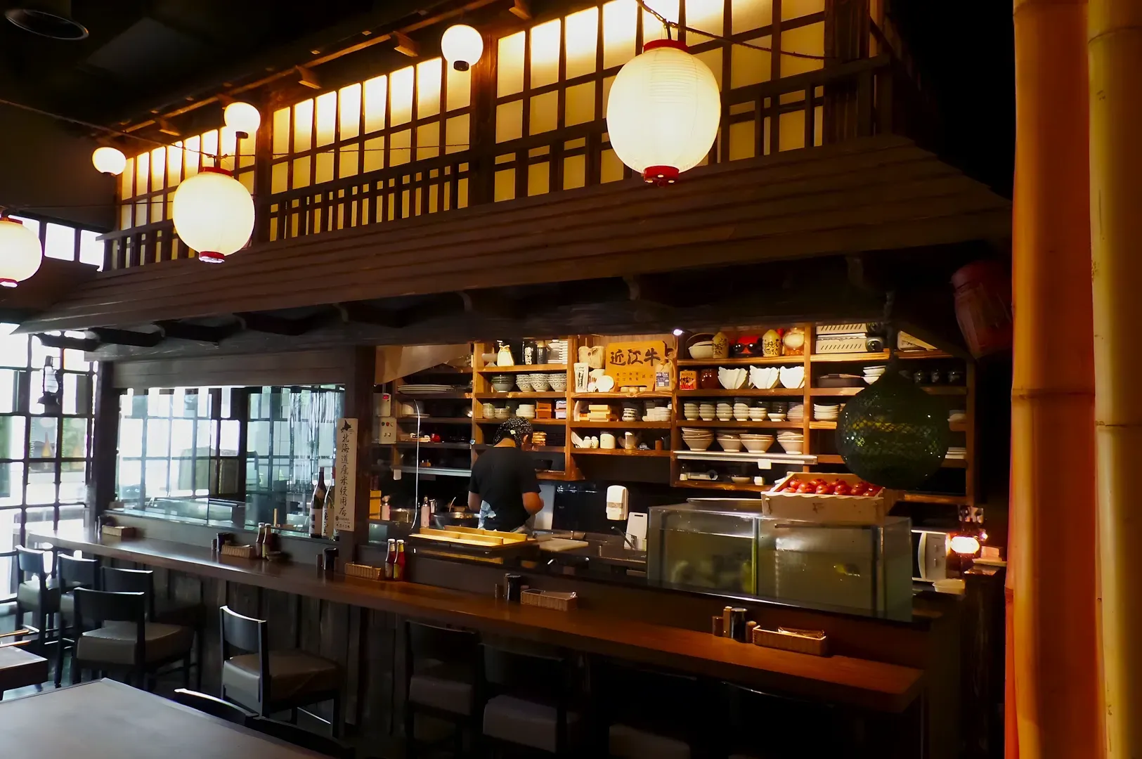 Cozy Japanese restaurant with warm lighting, lanterns, and wooden decor. A chef works behind the counter, surrounded by shelves of bowls and utensils.