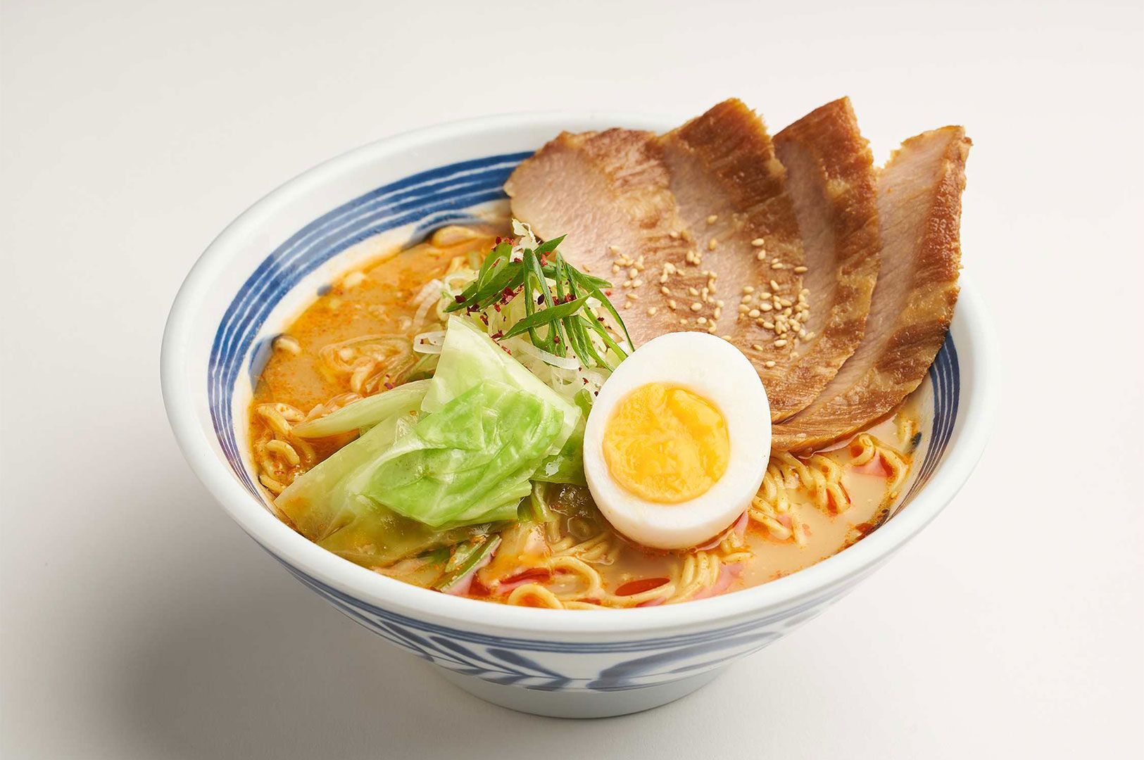 A bowl of rich miso ramen topped with sliced pork chashu, a soft-boiled egg, shredded scallions, and a piece of cabbage on a white background.