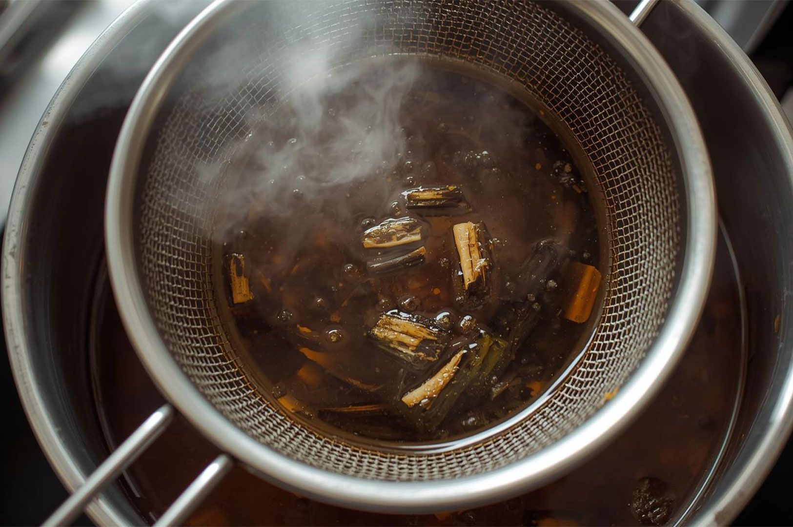 Overhead shot of steaming ramen stock being strained through a metal mesh sieve, showing pieces of kombu (seaweed) used for dashi broth.
