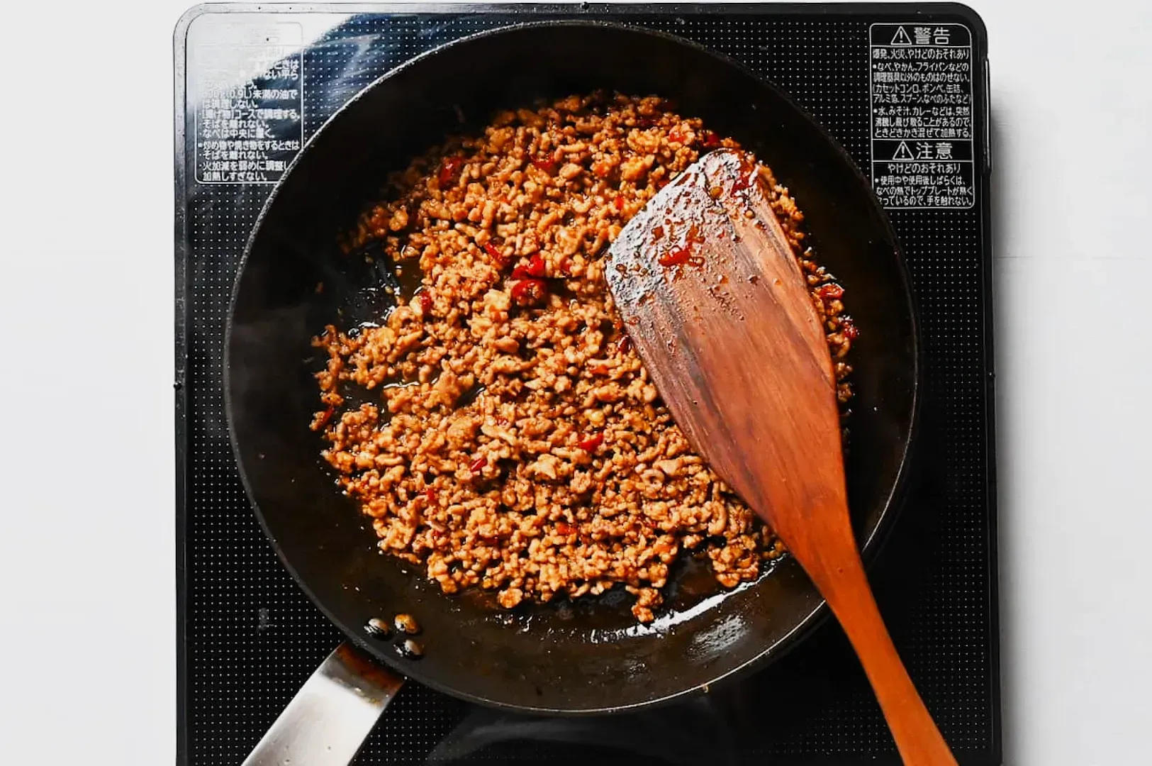 A pan filled with sizzling browned ground meat mixed with bits of red pepper, placed on a stove. A wooden spatula rests on the pan, conveying a cooking process.