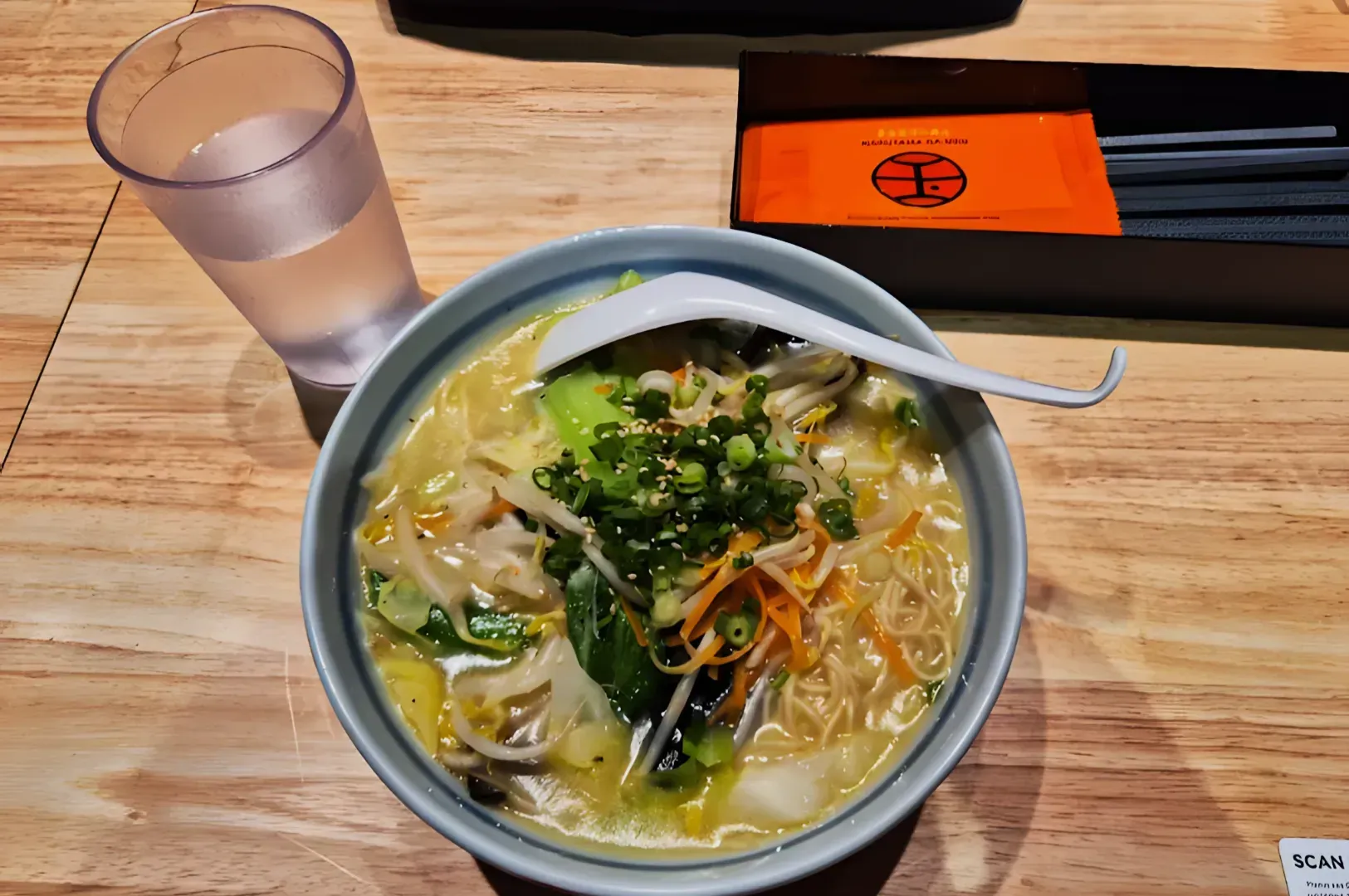 A bowl of ramen with vegetables, green onions, and a white spoon is on a wooden table. A glass of water and chopsticks are nearby, creating a cozy, inviting scene.