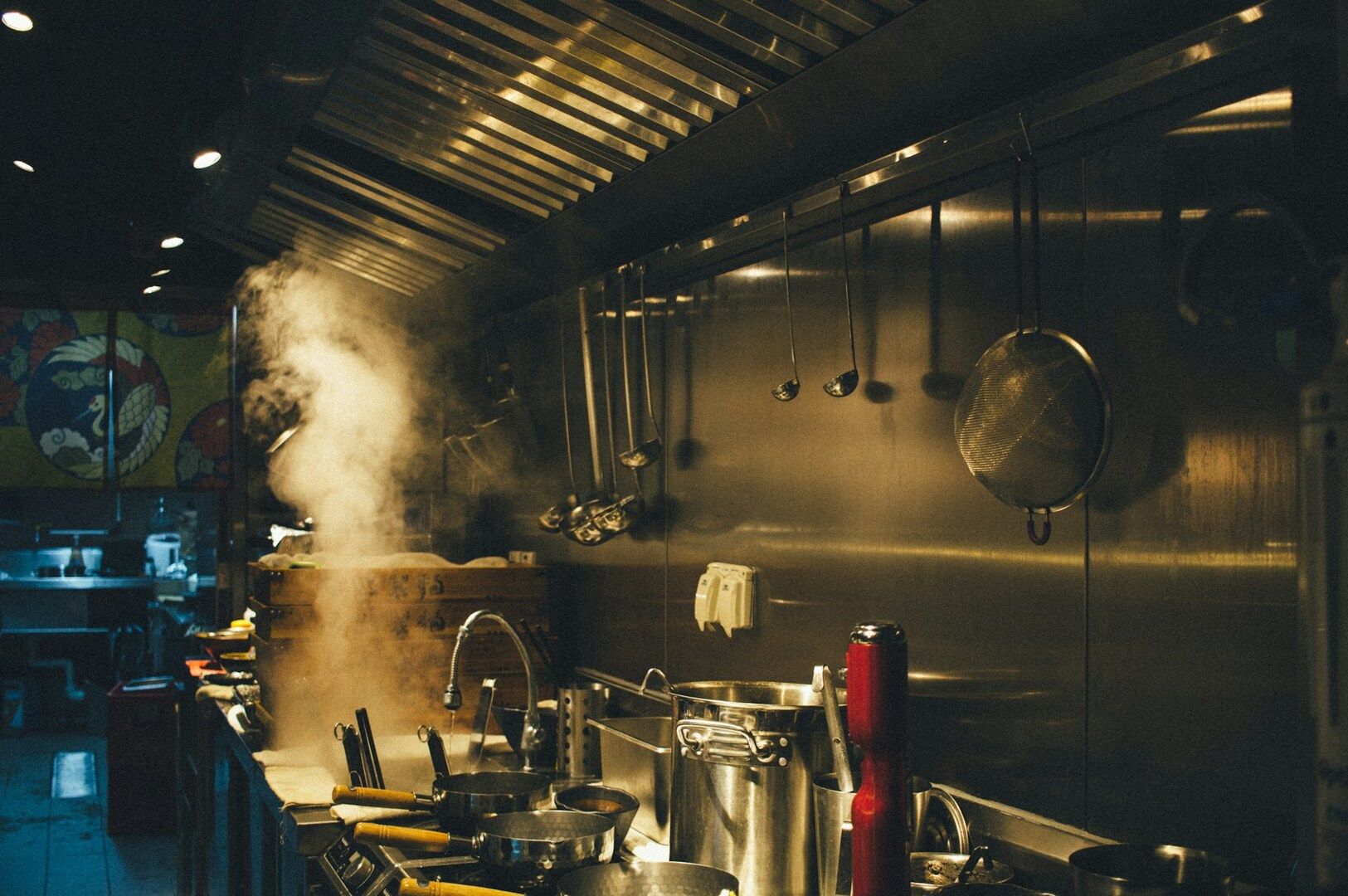 Commercial kitchen with steam rising from a pot, utensils hanging on a metallic wall, creating a warm and busy atmosphere. Cooking activity is evident.
