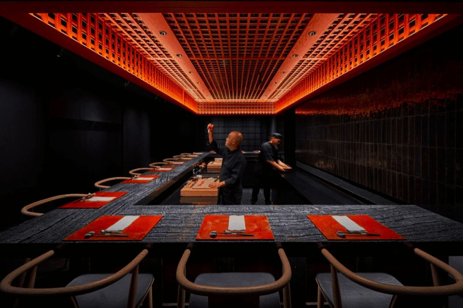 Sleek modern sushi restaurant with black walls and orange-lit ceiling. Chefs in black prepare sushi behind a U-shaped counter set with red placemats.
