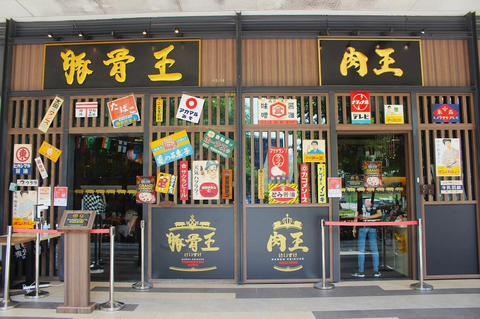 Exterior view of a Japanese restaurant with dark wooden lattice panels, gold lettering on black signs, and a window display crowded with colorful vintage posters, banners, and decals covering the glass.