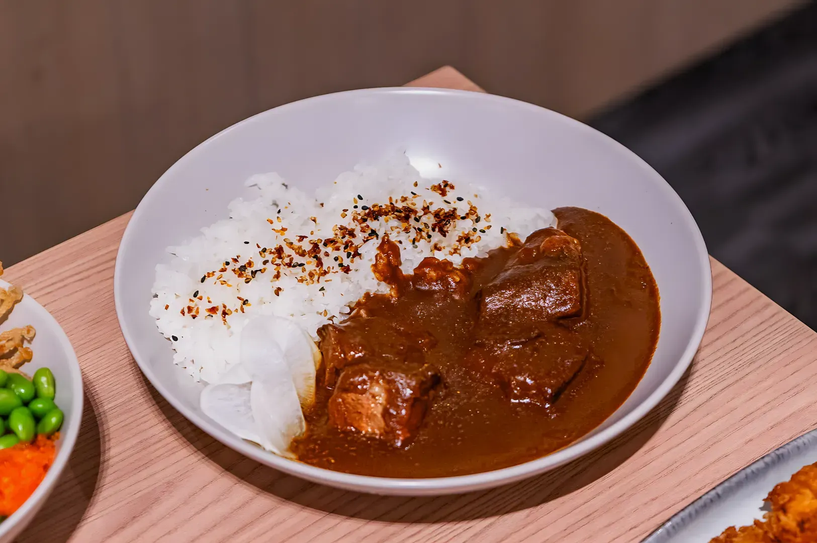 A white bowl on a wooden table contains rice garnished with spices, beef curry with chunks, and pickled radish slices, conveying a warm, inviting meal.