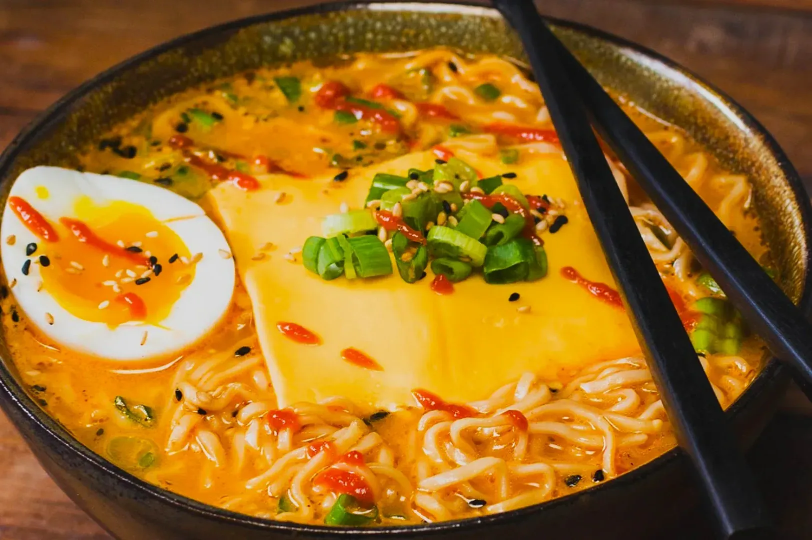 A bowl of ramen noodles in a rich, orange broth topped with a soft-boiled egg, melted cheese, chopped green onions, and sesame seeds, with chopsticks on the side.