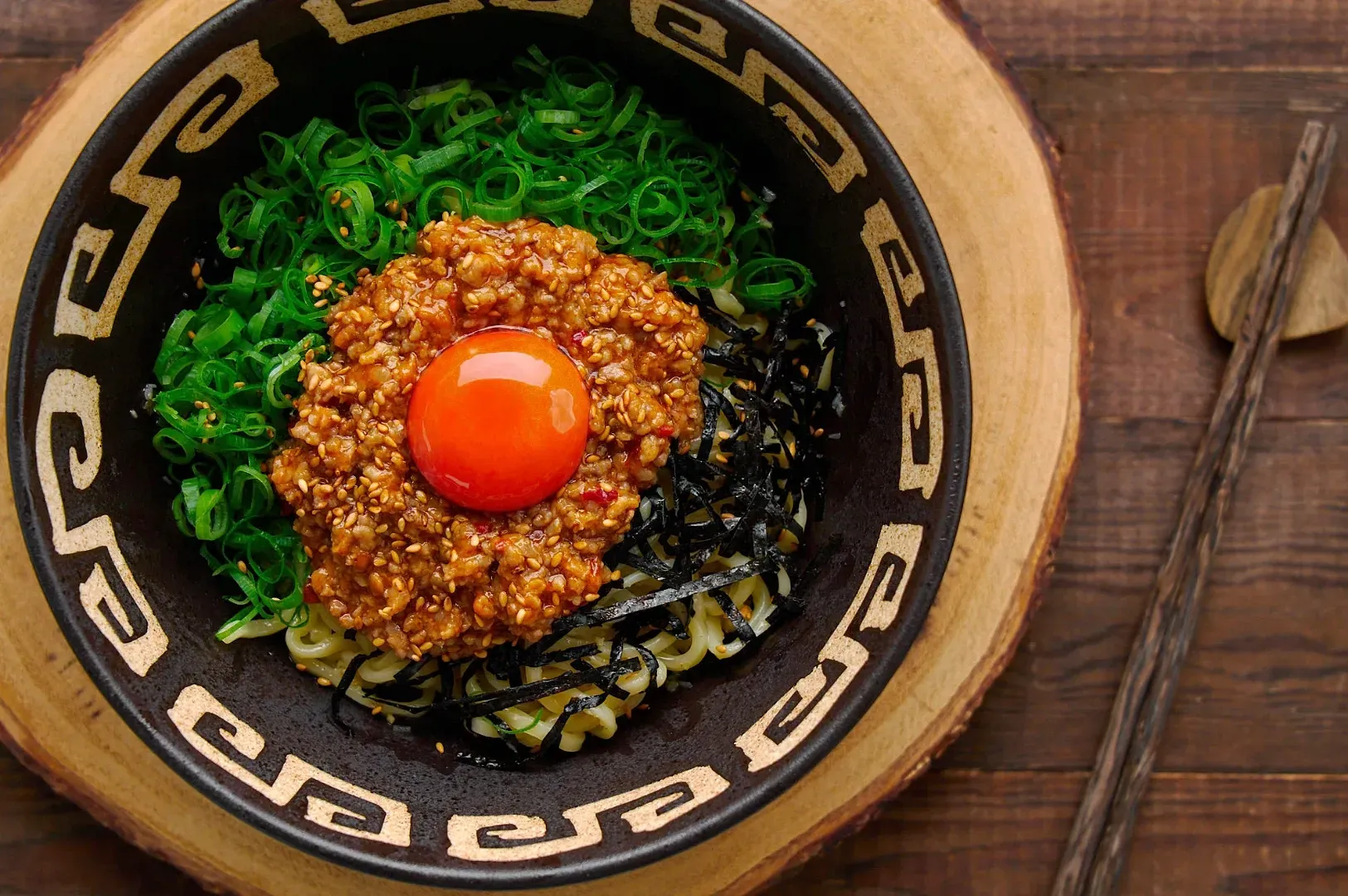 A bowl of ramen topped with a vibrant orange egg yolk, minced meat, green onions, and seaweed, placed on a wooden surface with chopsticks nearby.