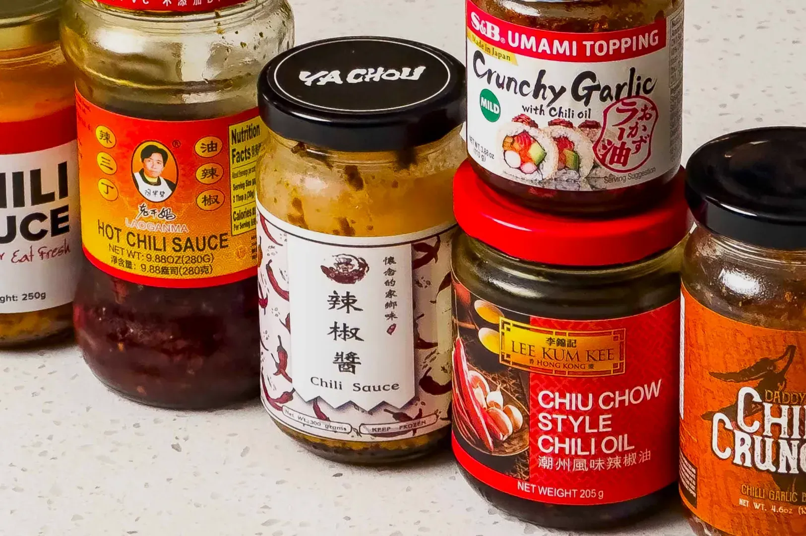 A collection of various chili sauces and oils in jars on a counter. Labels feature vibrant designs and text in different languages, evoking spicy flavors.