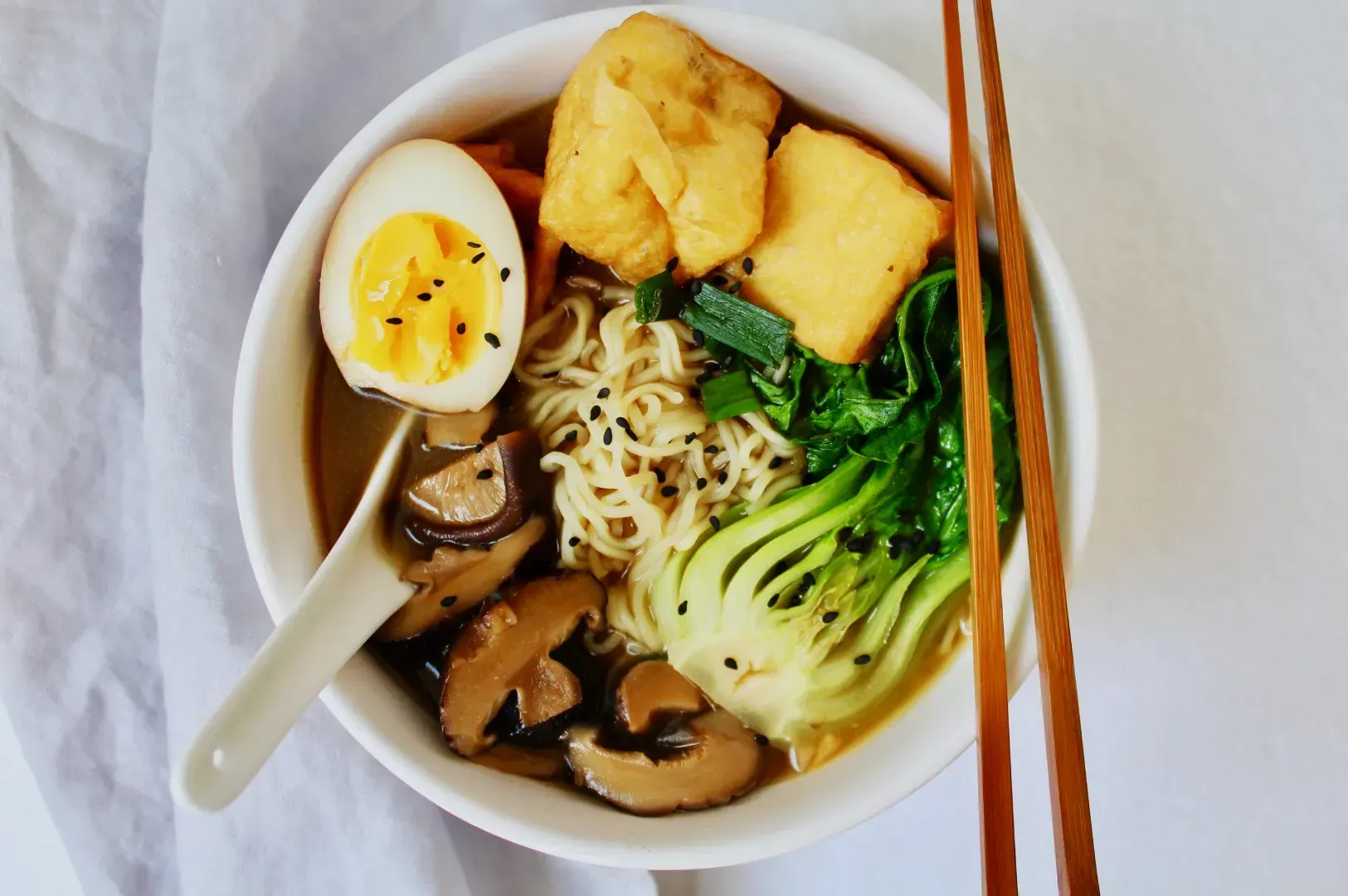 A bowl of ramen topped with bok choy, tofu, mushrooms, half a soft-boiled egg, and sesame seeds. Chopsticks rest on the rim, conveying a savory, comforting meal.