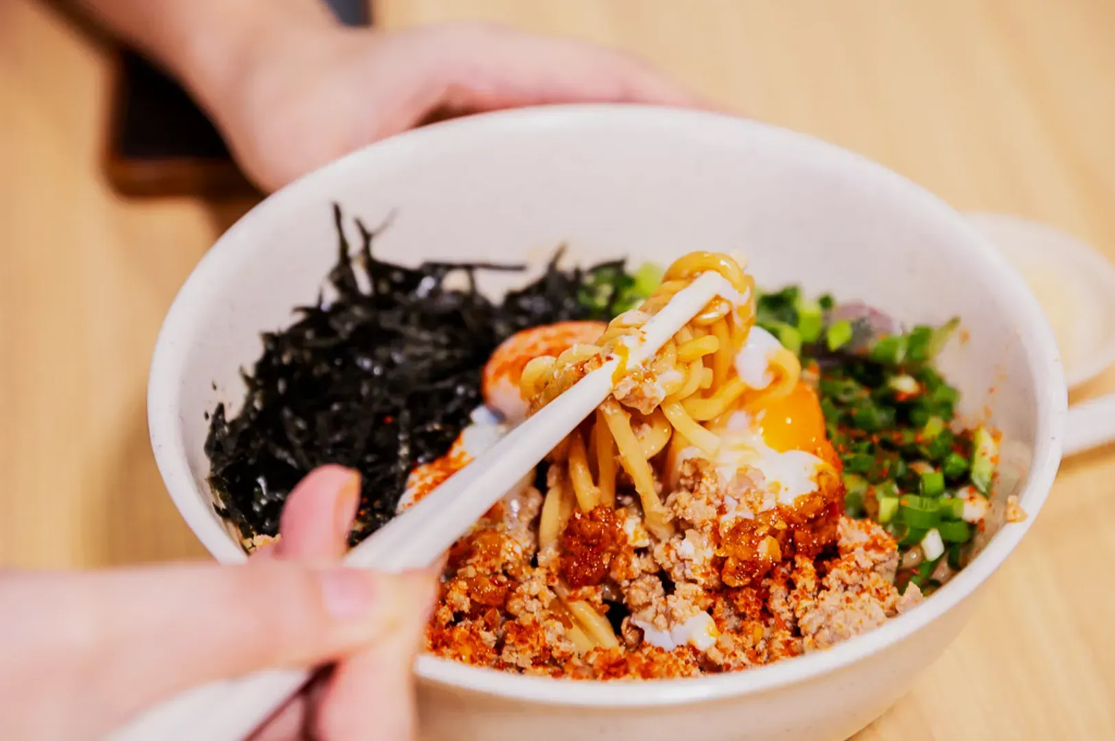 A bowl of noodle dish topped with minced meat, seaweed, chopped green onions, and an egg yolk. Chopsticks hold some noodles, conveying a savory appeal.