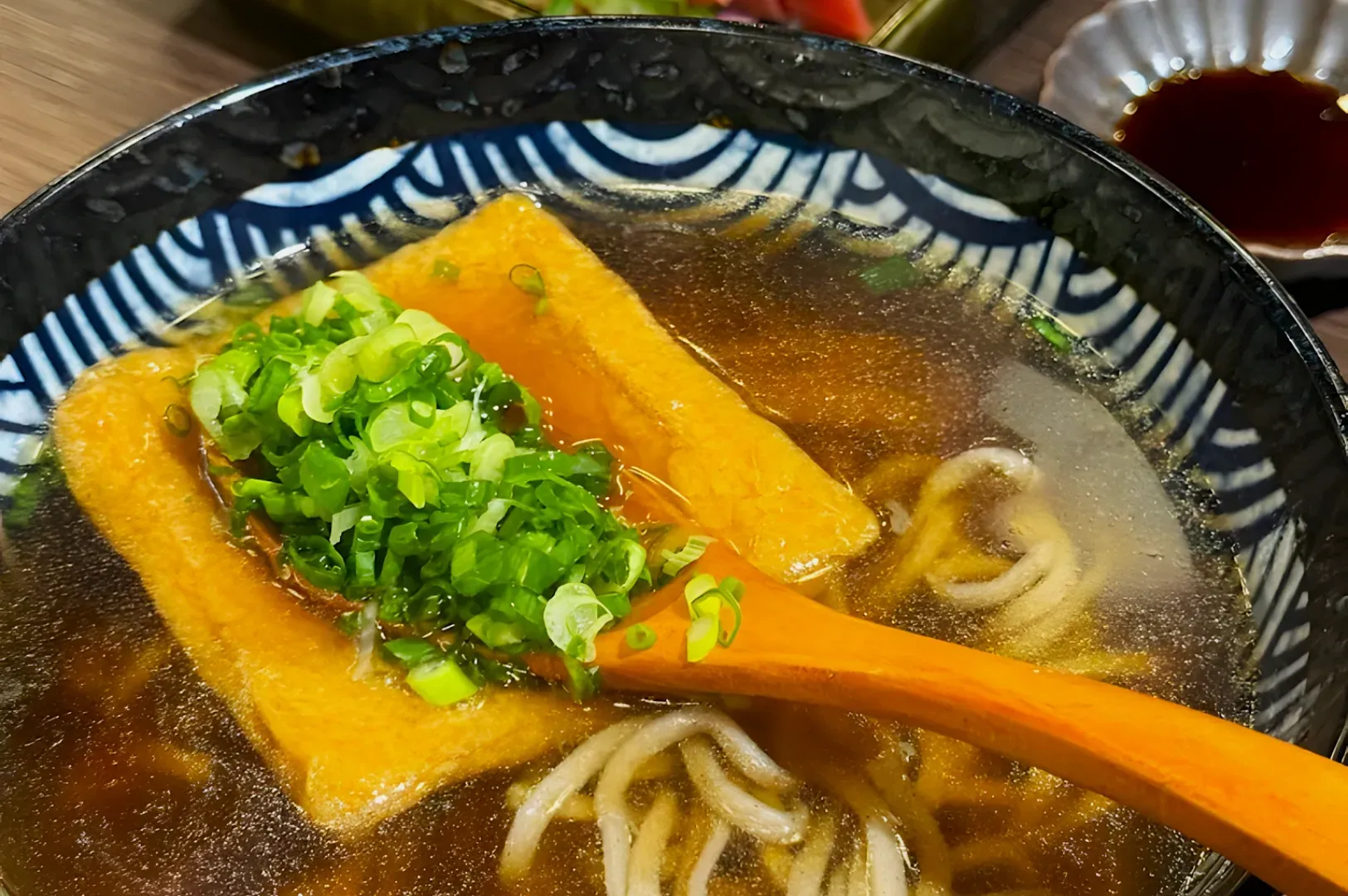 Close-up of a bowl of udon soup with a light broth. A wooden spoon holds sliced green onions and fried tofu, enhancing the enticing presentation.