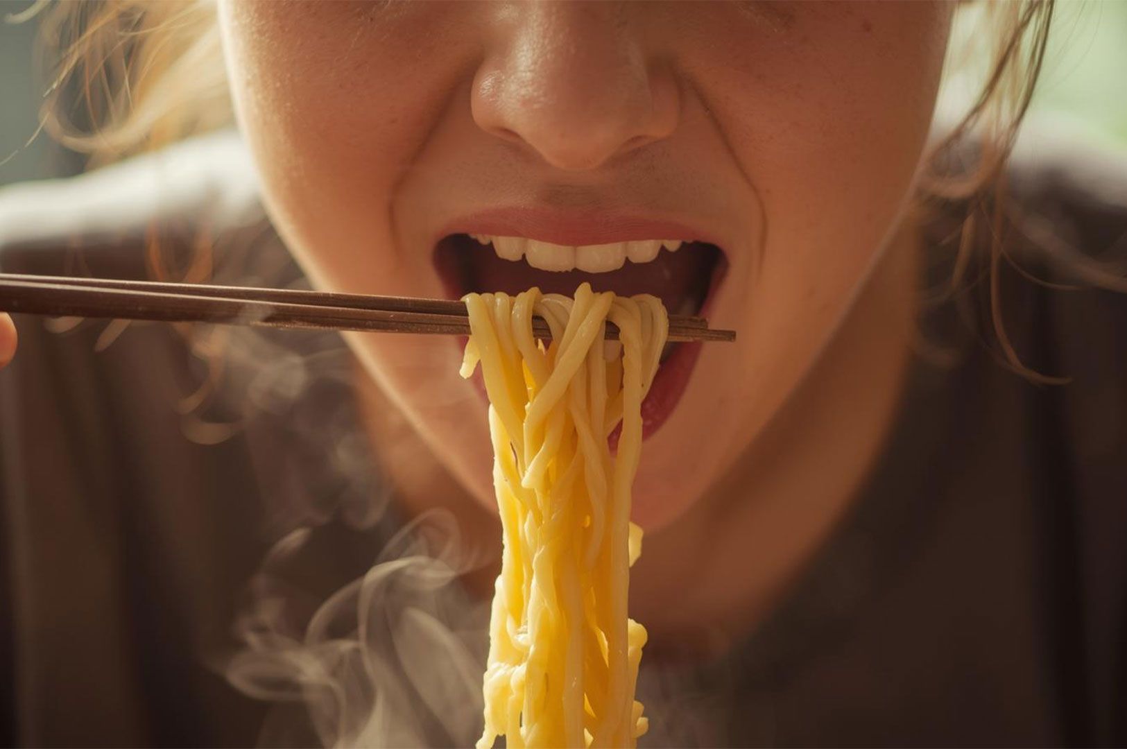 An extreme close-up of a person's mouth and lower face, demonstrating the enjoyment of a hot meal by slurping a large strand of steaming ramen noodles held by chopsticks.