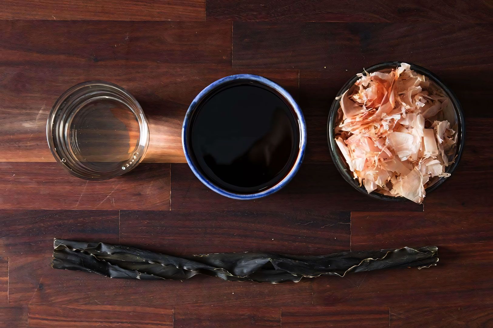 Overhead flat-lay of ramen broth ingredients on a dark wood surface: a long strip of dried kelp (kombu), cups of mirin, dark soy sauce (shoyu), and a bowl of bonito flakes.