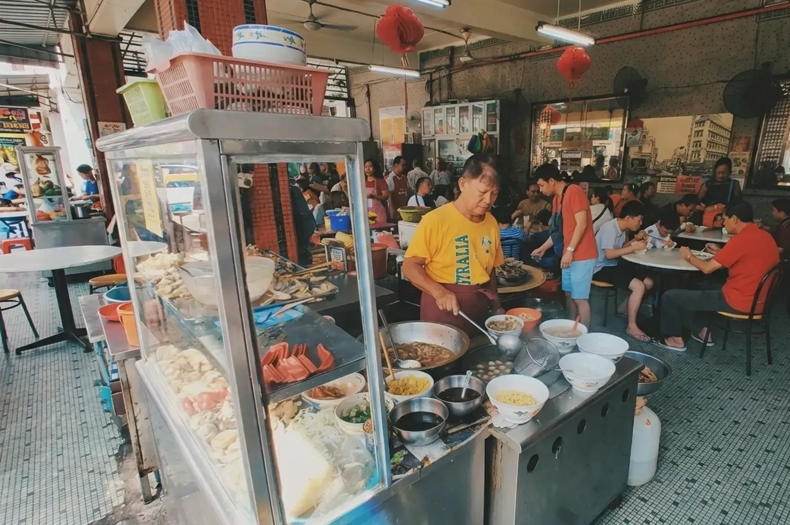 Busy Singaporean food stall vendor preparing ramen or noodle soup at hawker center with customers dining in background.