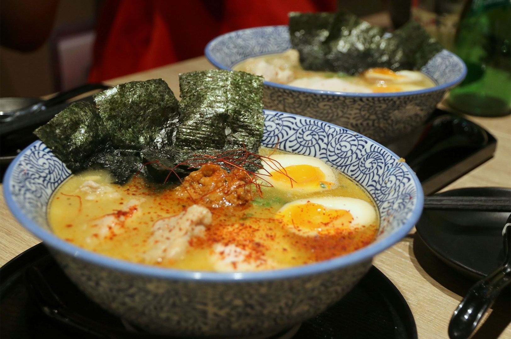 A close-up of two bowls of rich, orange-tinted spicy ramen soup, topped with half soft-boiled eggs, crumbled ground meat, chili flakes, and large sheets of toasted nori seaweed.