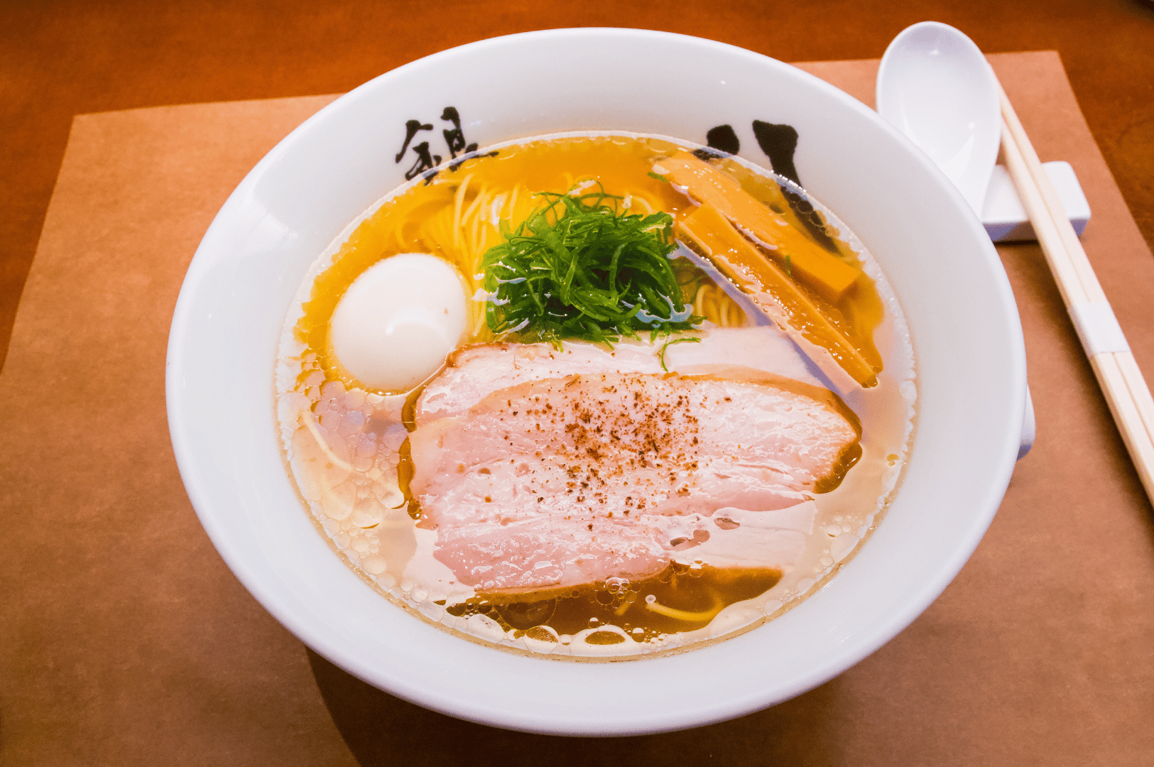 A cleanly presented bowl of ramen served in a white bowl with Japanese characters on the rim, featuring clear broth, soft-boiled egg, peppered pork belly slices, bamboo shoots, green onions, and noodles. A white spoon and wooden chopsticks rest on a napkin beside the bowl, all set on a brown surface. The minimalist composition and refined ingredients reflect Singapore’s appreciation for traditional Japanese ramen with modern precision.