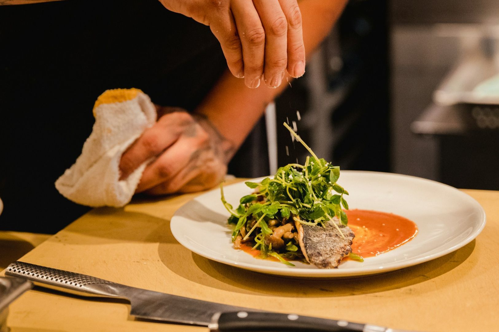A chef sprinkles salt on a gourmet dish with greens and seared fish, elegantly plated on a white dish. A focused culinary creation moment.
